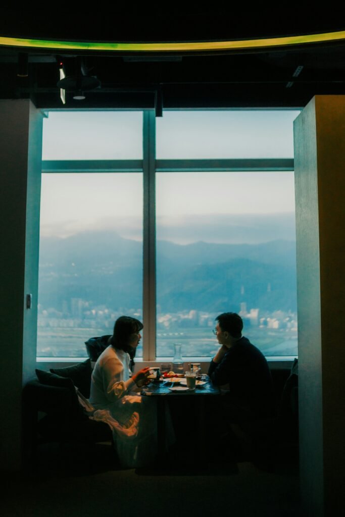 Two people sit across a small dining table beside a large window overlooking the city.