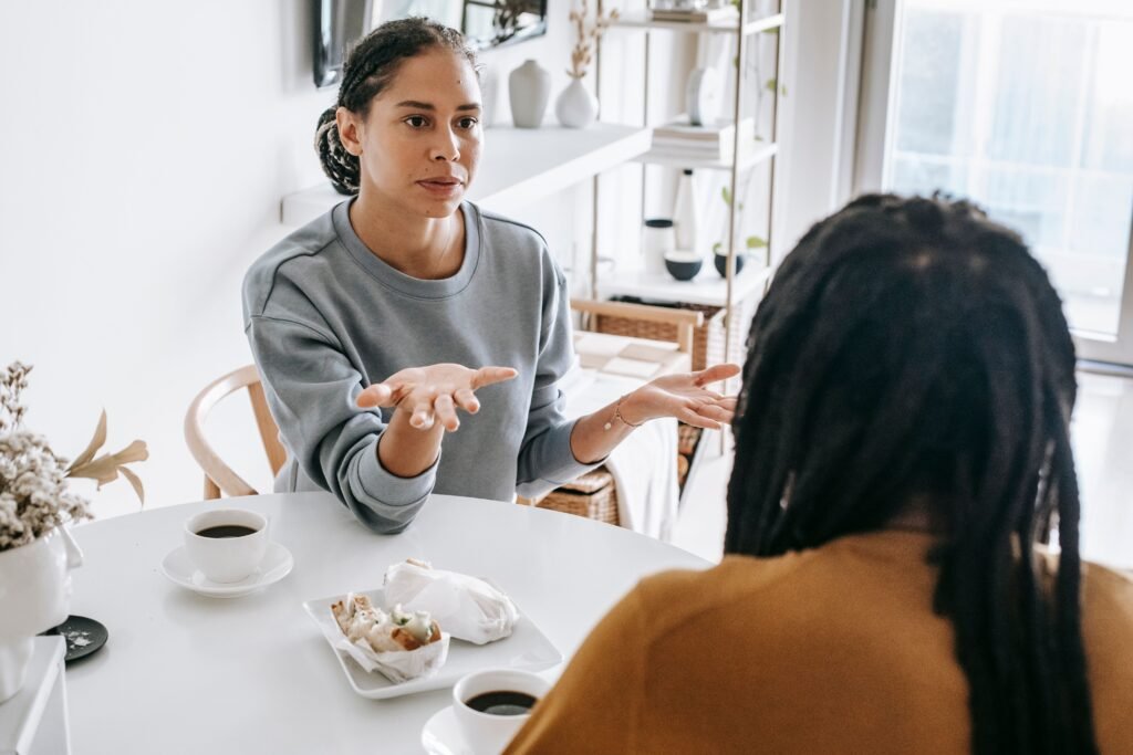 A woman gestures with open palms while talking to a man across a table with coffee cups.