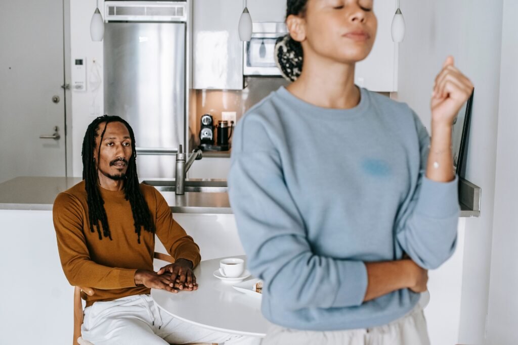 Woman standing with her arms crossed while a man sits at a kitchen table behind her.