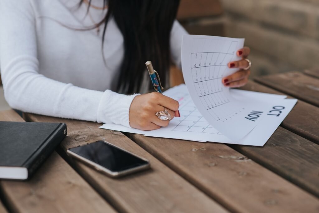 Woman writing on a paper calendar at a wooden table, with a smartphone and notebook nearby.