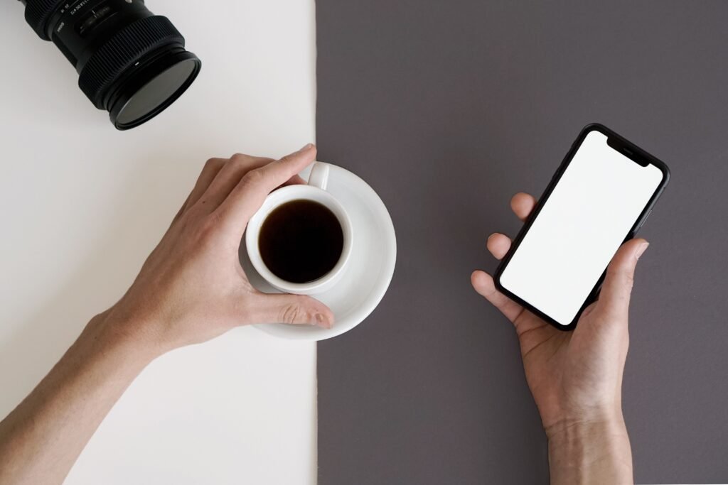 Person holding a white coffee cup and a smartphone with a blank screen at a table.