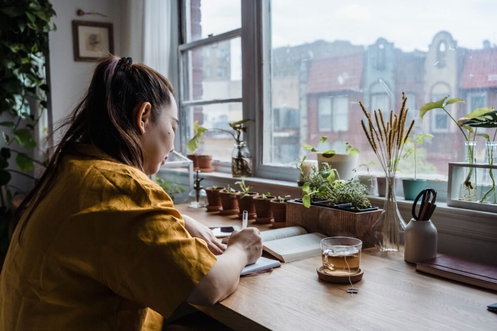 A woman writes in a notebook beside a window lined with plants.