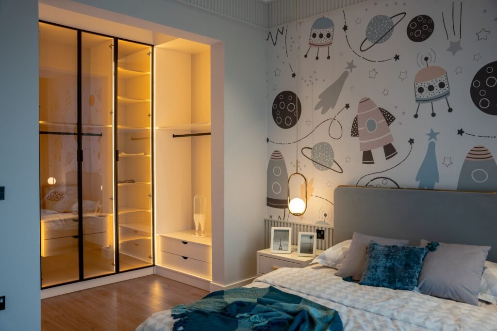 An empty child's bedroom with space-themed wall art, a made bed, and a glowing bedside lamp.