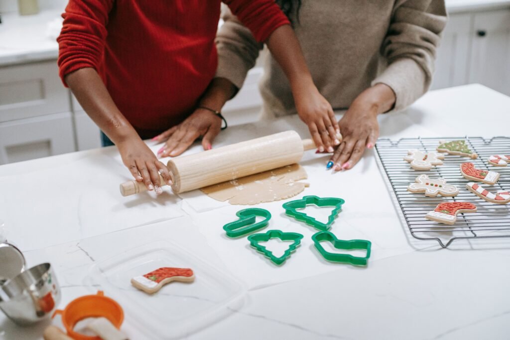 Two people roll dough together beside holiday cookie cutters and decorated cookies.