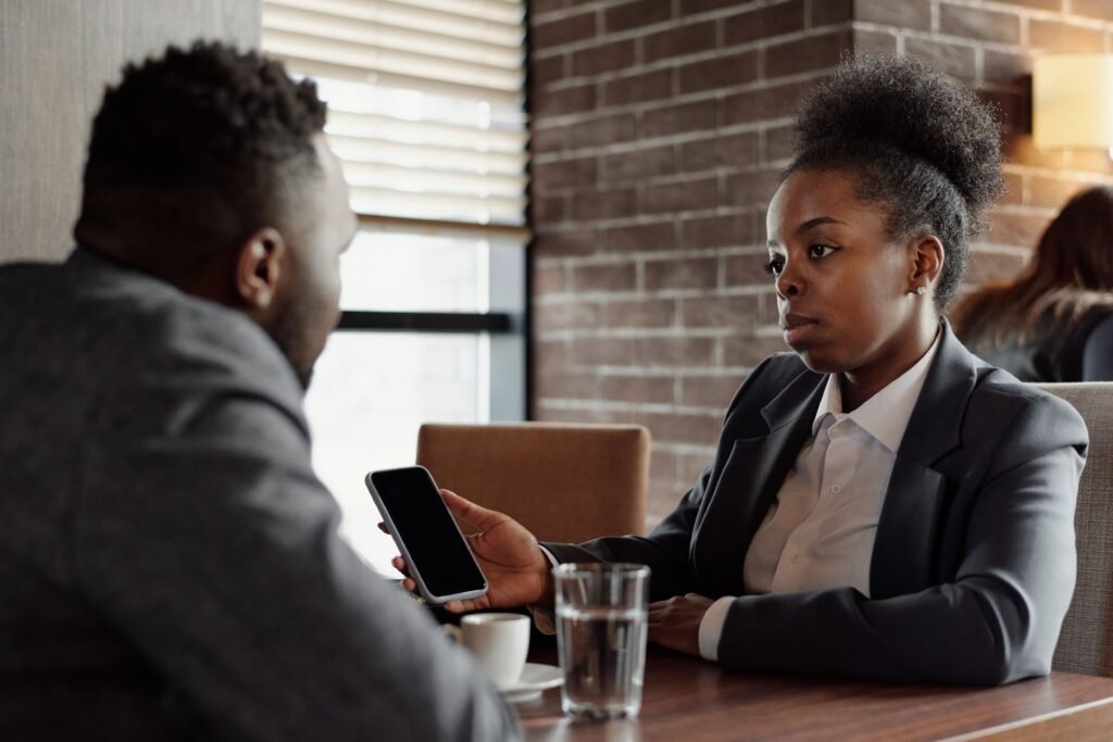 Woman holding up a smartphone while speaking with a man across a table.