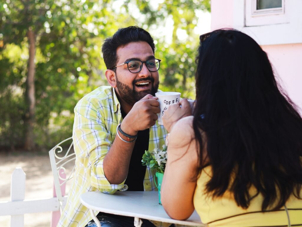 Man and woman sitting across from each other at an outdoor café table, smiling and enjoying a conversation over coffee.