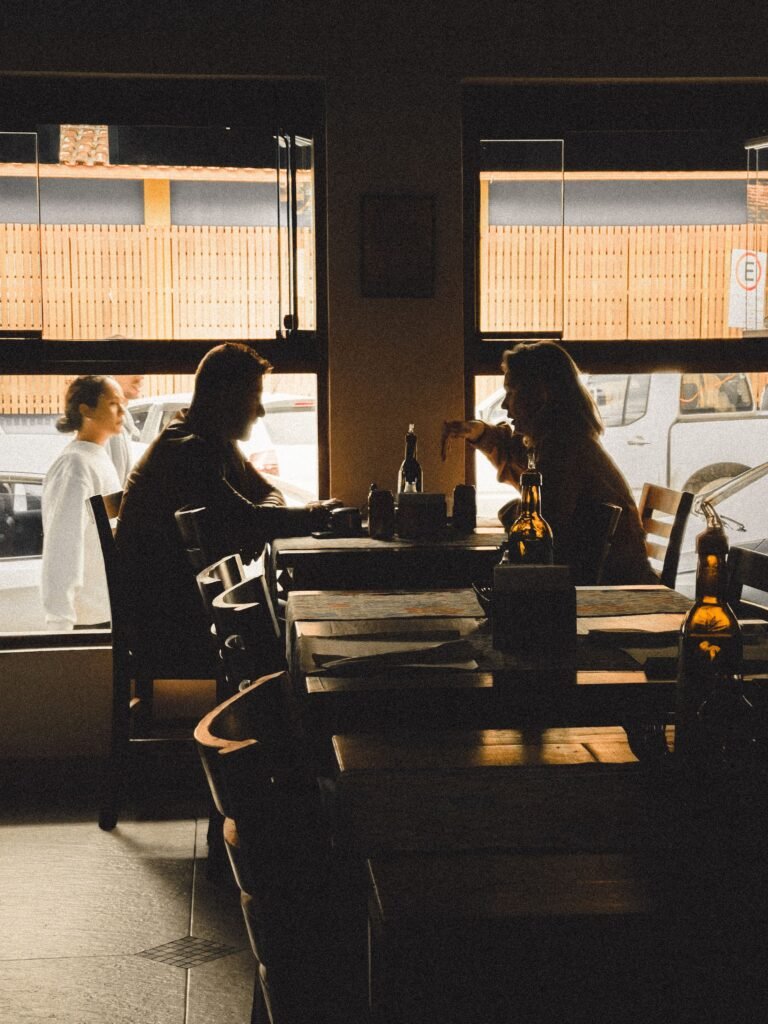 Two people sitting across from each other at a table in a dimly lit café, having a conversation near the window.