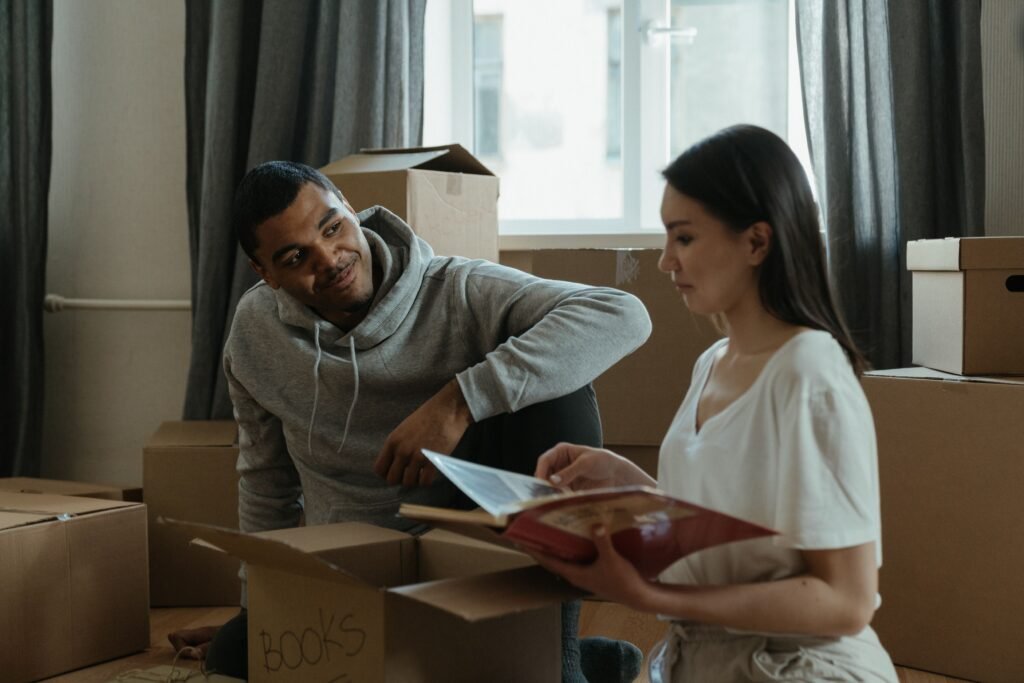 Woman holding an open photo album while a man sits beside her among cardboard moving boxes.