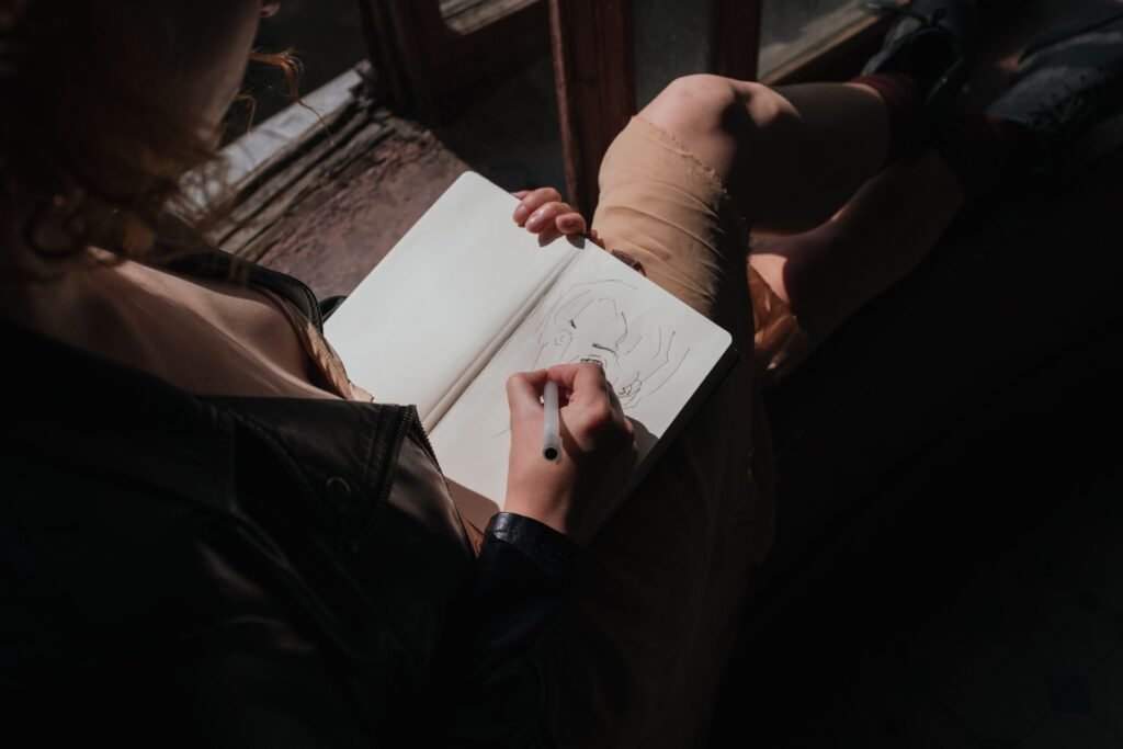 Person sitting by a window, drawing a portrait in a sketchbook with a pen.