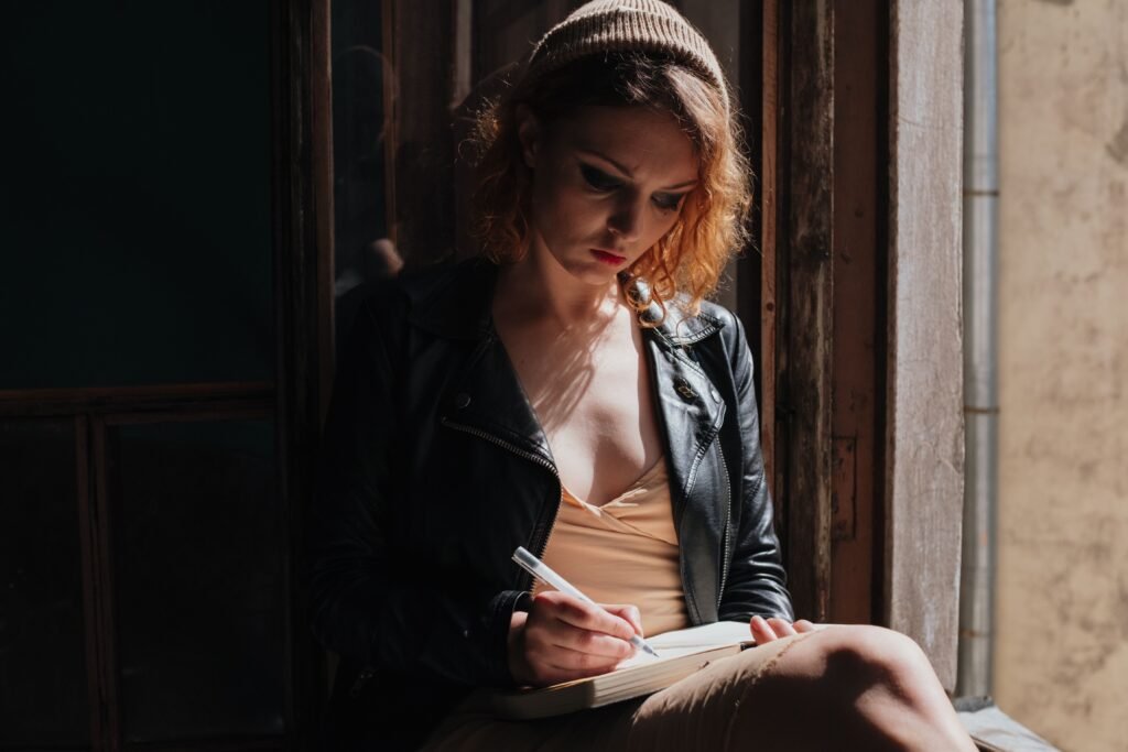 Woman in a knit hat and leather jacket writing in a notebook beside a sunlit window.
