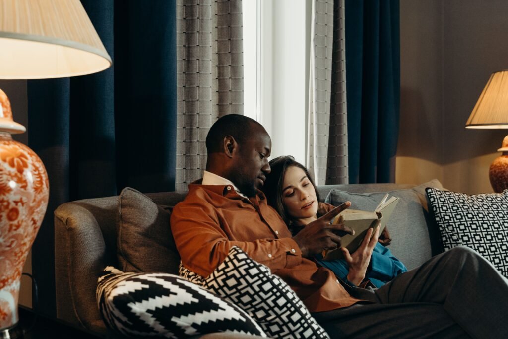 Man and woman reading a book together on a sofa, with the woman resting against him as he points to a page.