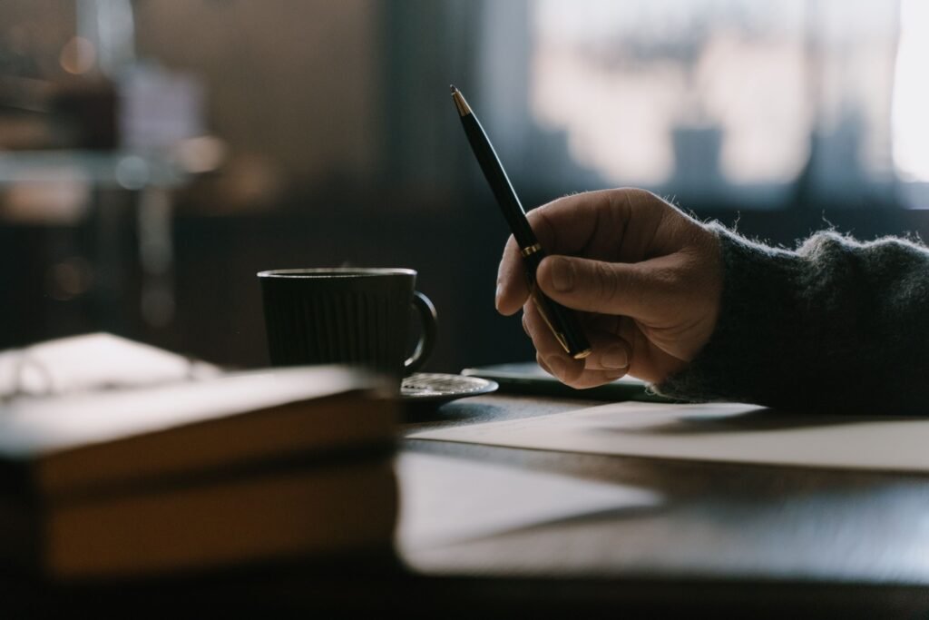 Close-up of a person holding a pen over paper beside a cup of coffee, preparing to write notes at a wooden table.