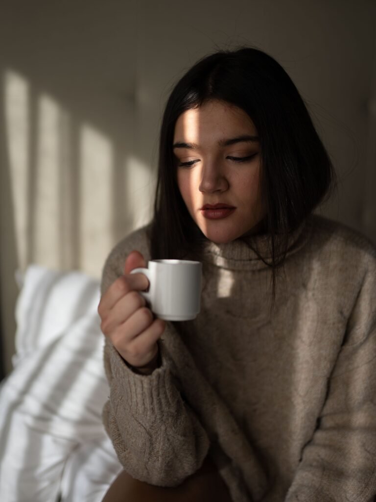 Thoughtful woman sitting alone indoors, holding a white coffee cup and looking down as soft sunlight falls across her face.