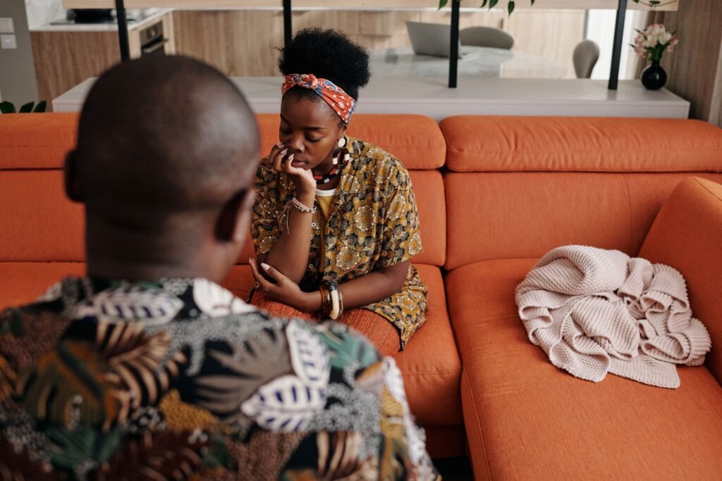 A woman sits with her hand against her face while another person faces her across the room.
