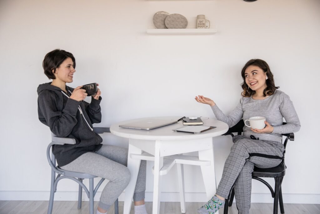 Two women smile and talk while holding mugs at a round white table with a laptop, tablet, and notebook.