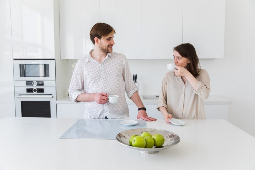 A man and woman holding cups and looking at each other in a kitchen.