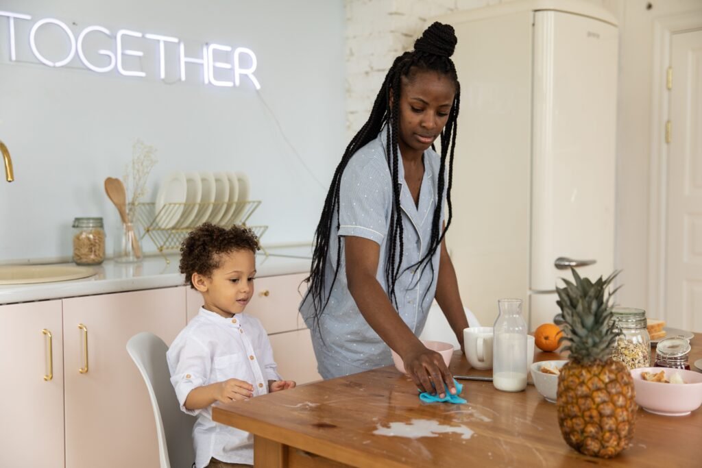 Woman wiping a kitchen table while a child stands beside her.