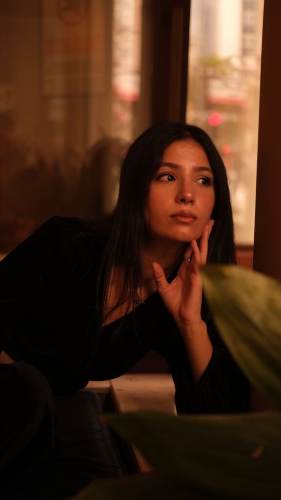 A woman sits beside a window in warm light, resting her hand against her cheek and looking to the side.