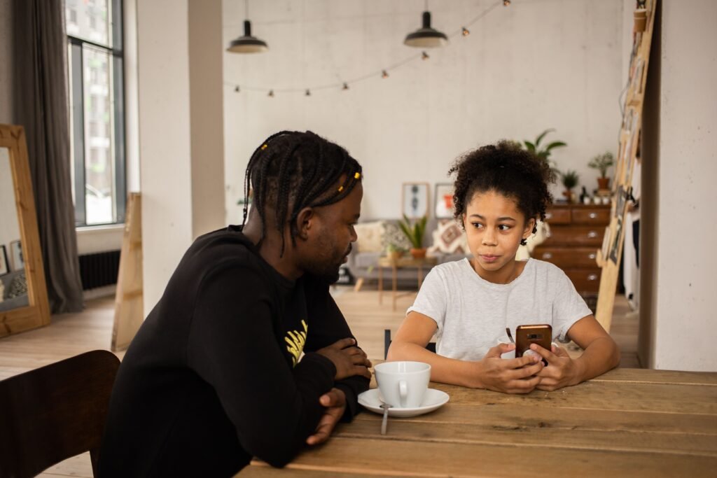 Man leaning toward a girl holding a smartphone as they sit together at a wooden table.