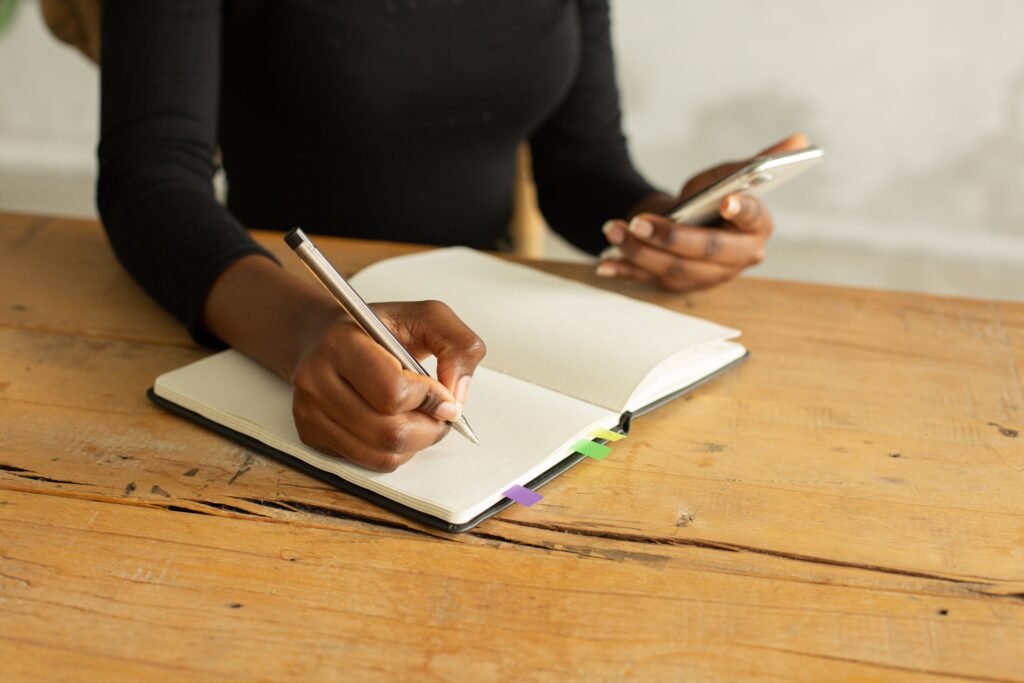 Person writing in an open notebook while holding a smartphone at a wooden table.