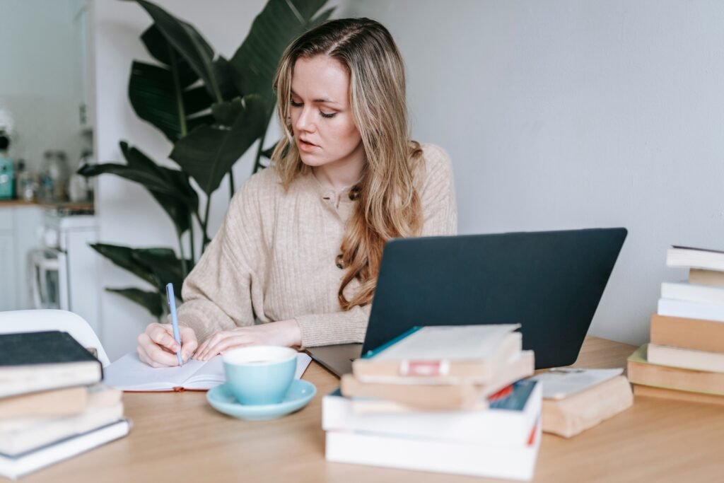 A woman writes in a notebook beside an open laptop, a blue cup, and stacks of books.