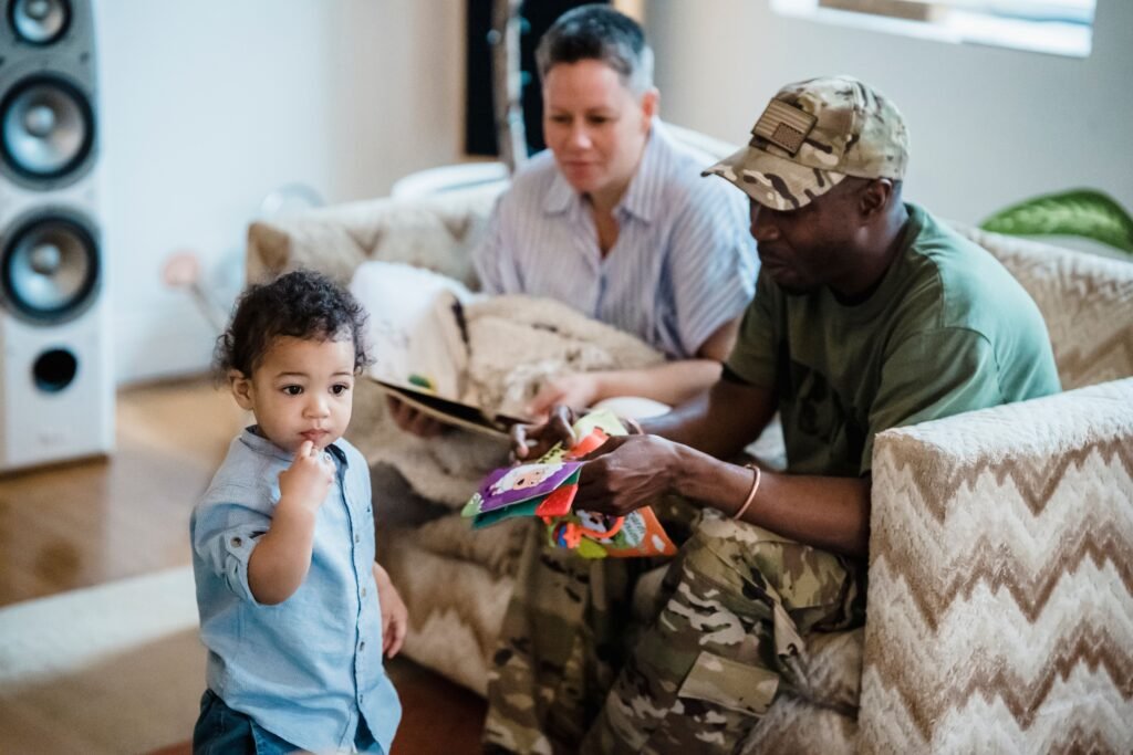A young child stands beside two seated men, one holding a colorful fabric activity book.