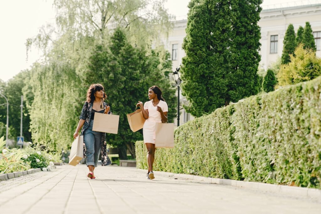 Two women carry shopping bags and talk while walking along a tree-lined path.
