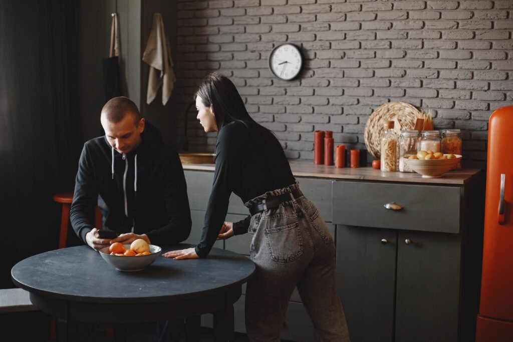 Man looking down at his smartphone while a woman leans toward him across a kitchen table, appearing to seek his attention.