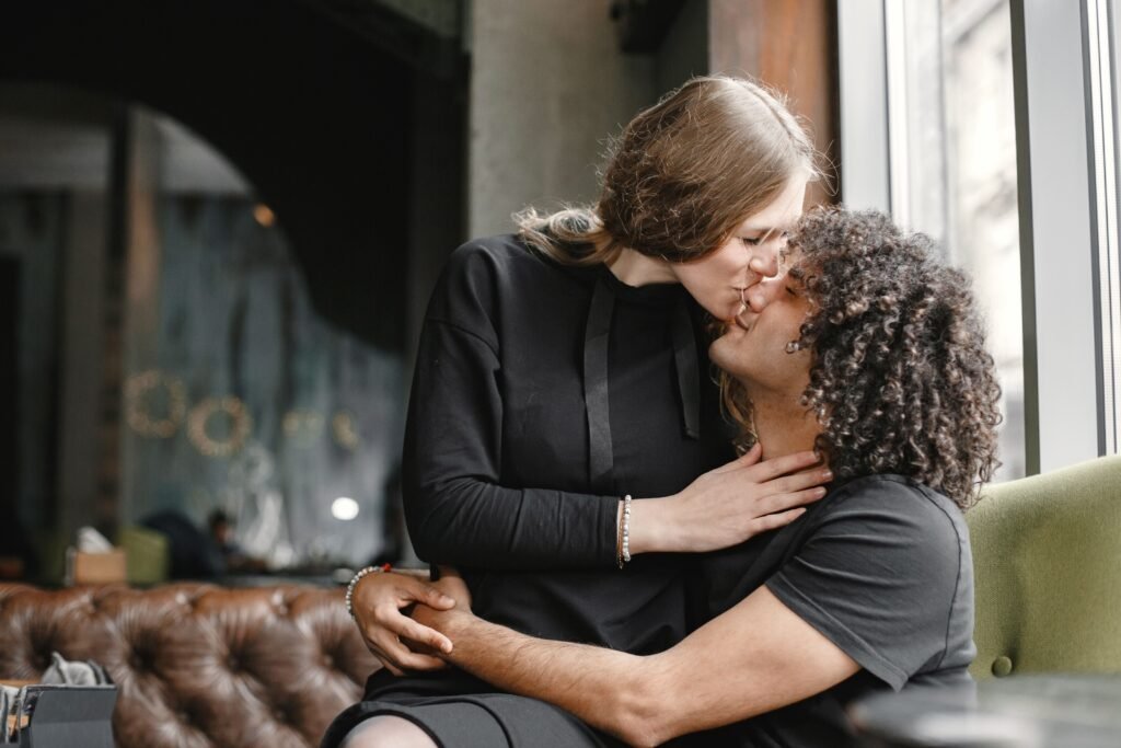 Woman leaning down to kiss a seated man beside a window as he holds her around the waist.