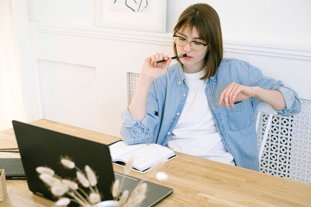 Woman wearing glasses holding a pen near her lips, with an open notebook and laptop on the table.