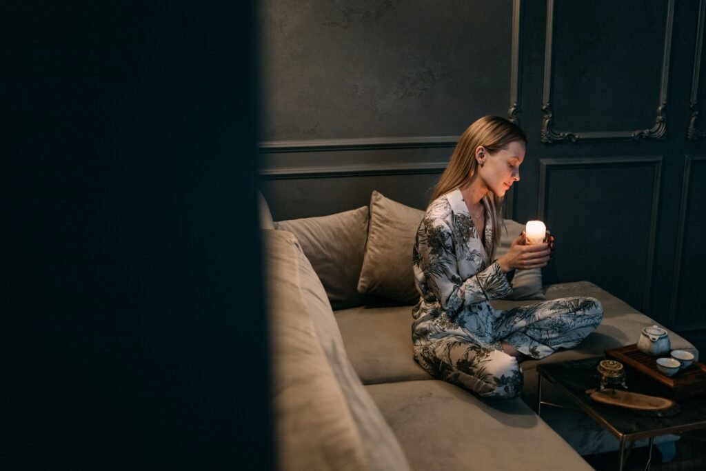 Woman sitting alone on a sofa in a dimly lit room, holding a glowing candle and enjoying a peaceful moment of reflection.