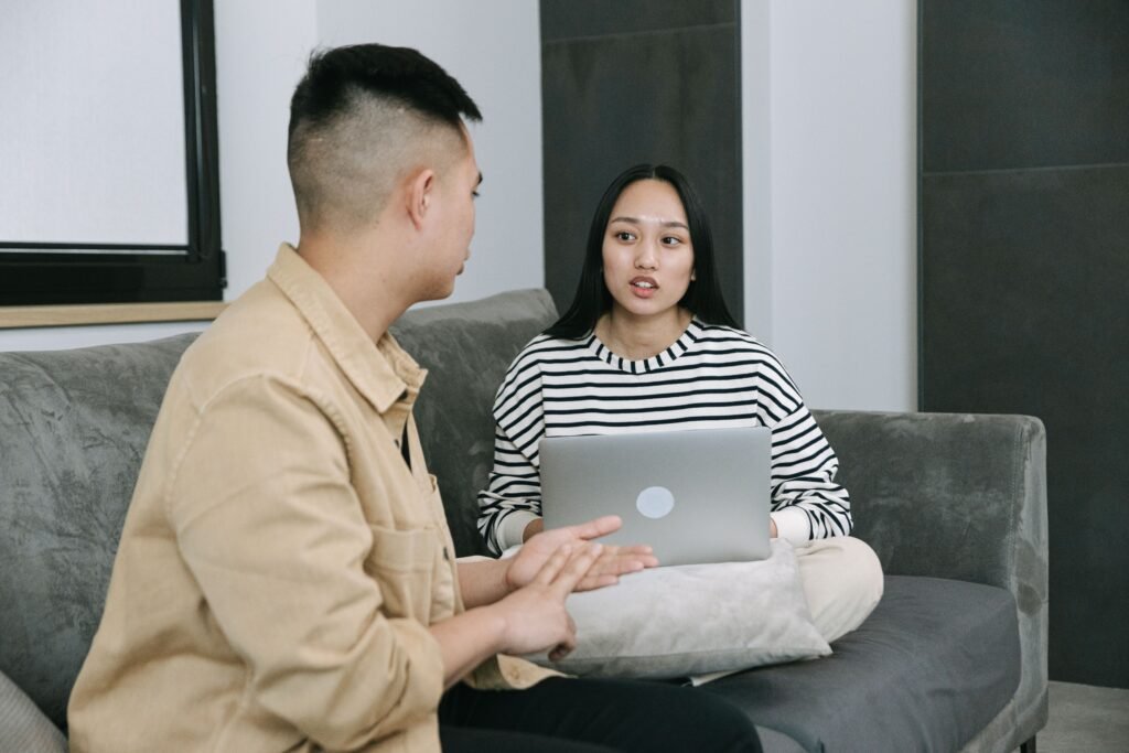 A man gestures during a conversation with a woman seated on a gray sofa with a laptop.