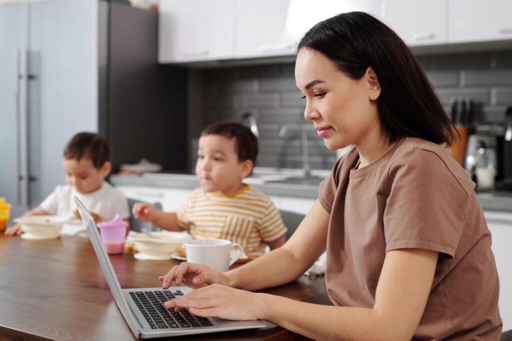 Woman typing on a laptop at a kitchen table while two children eat nearby
