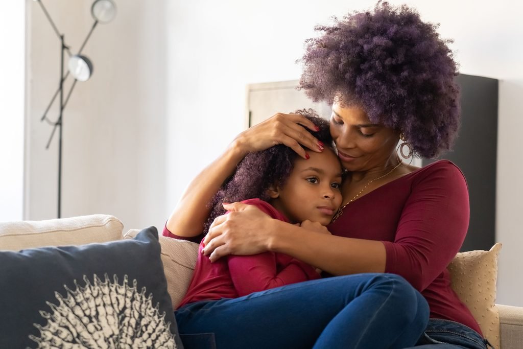 A woman holds a young girl close while they sit together on a sofa.