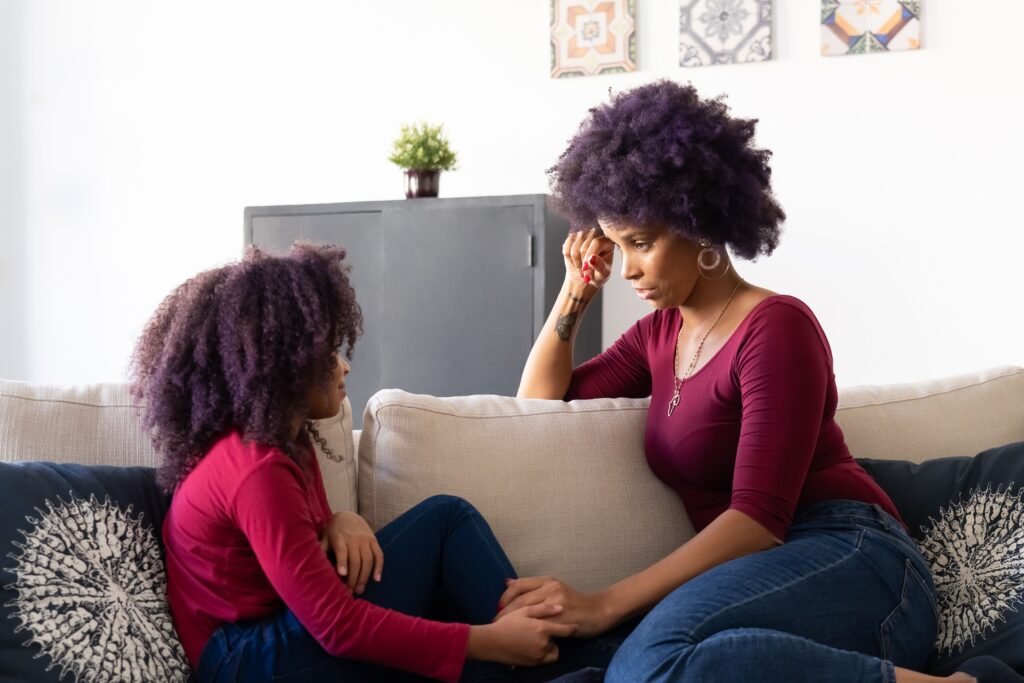 Woman and young girl sitting face to face on a sofa, holding hands while talking.
