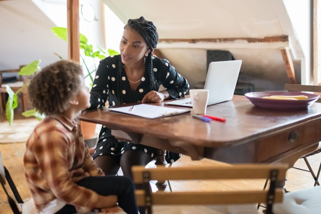 Woman listening to a child across a table with a laptop and notebook