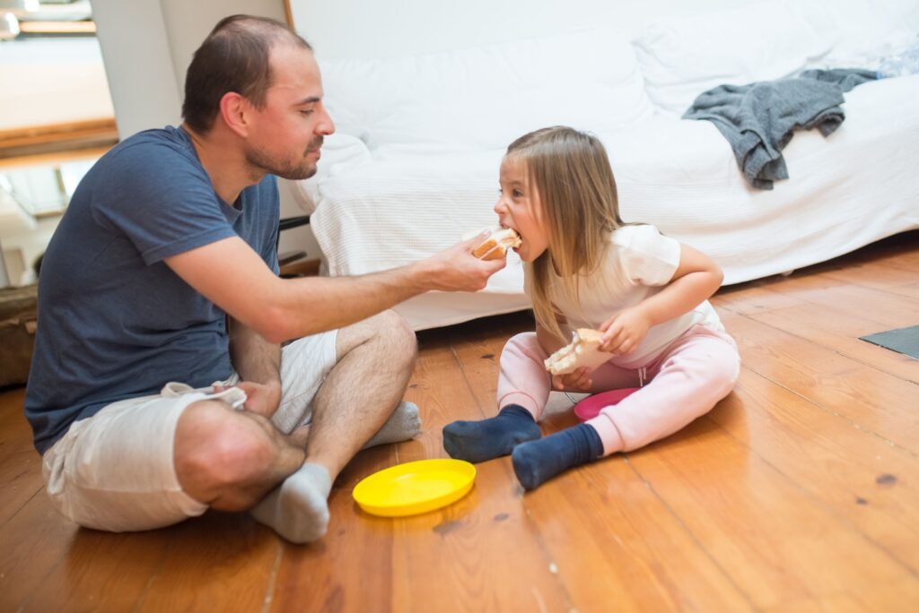 Man offers food to a young girl as they sit together on the floor at home.
