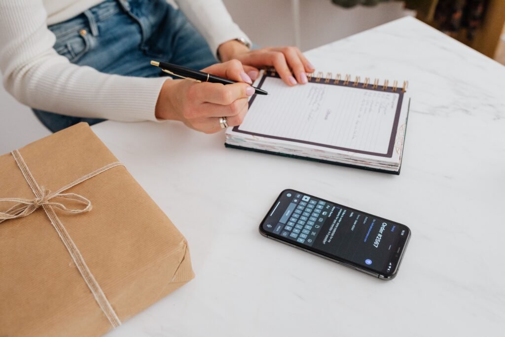 Person writing in a spiral notebook beside a smartphone and a parcel wrapped in brown paper.