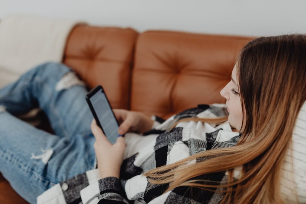 Woman relaxing on a brown leather sofa while reading messages on her smartphone at home.