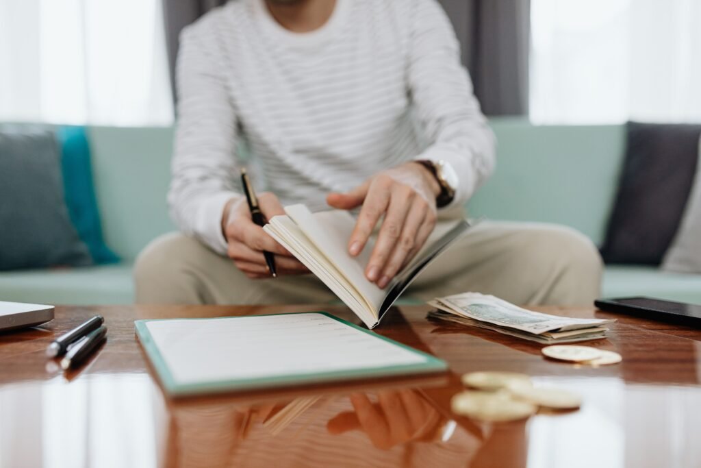 Person holding a pen and turning notebook pages beside cash, coins, and paperwork on a coffee table.