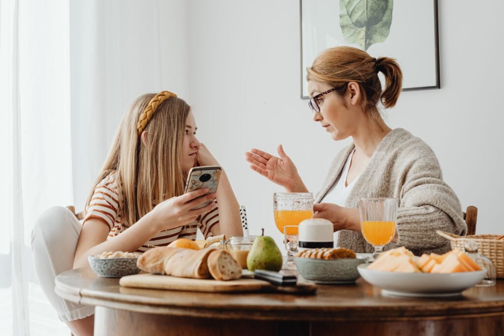 Woman gesturing while talking to a girl holding a smartphone at a breakfast table.