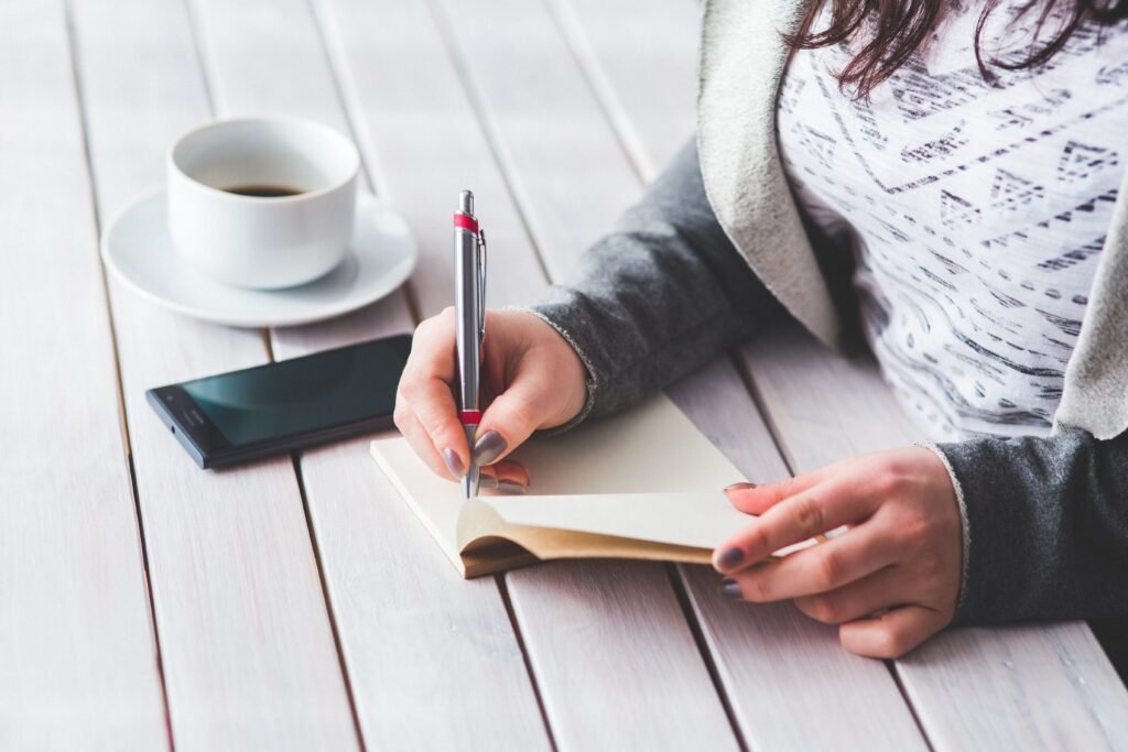 Person writing in a notebook beside a smartphone and a cup of coffee on a white wooden table.