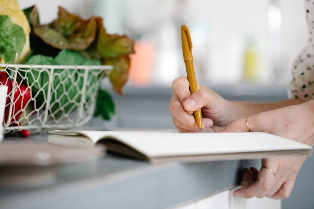 Close-up of a person writing in an open notebook beside a basket of vegetables.