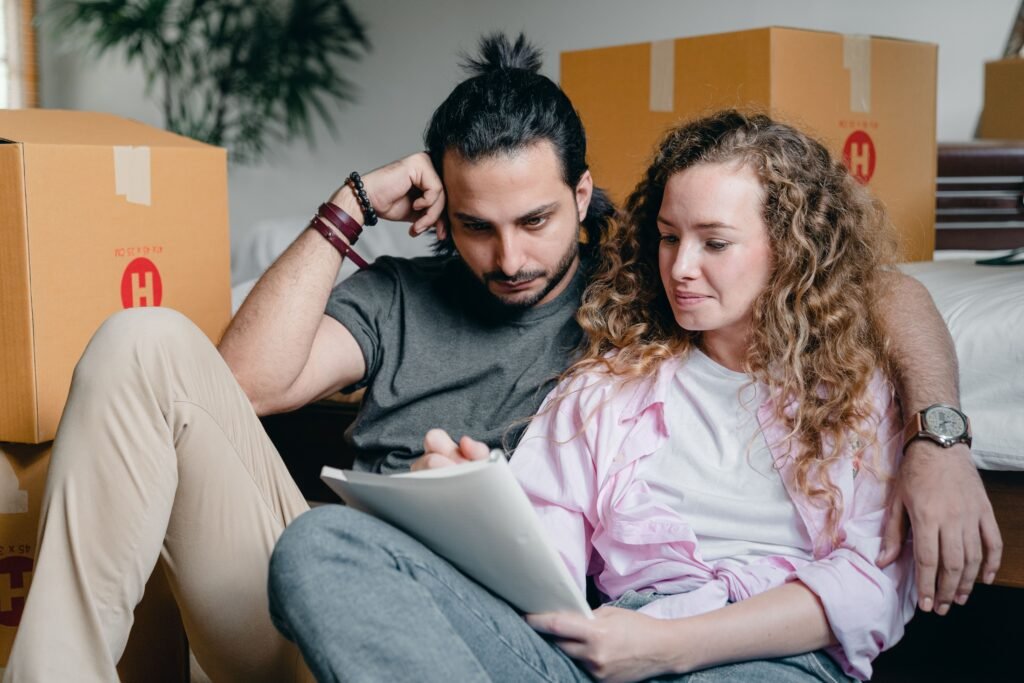 A couple sits among moving boxes and looks together at a notebook.