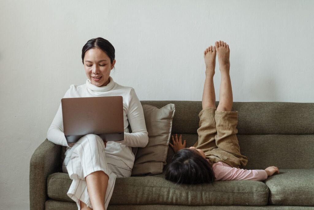 Mother working on a laptop while her young child plays beside her on the sofa at home.