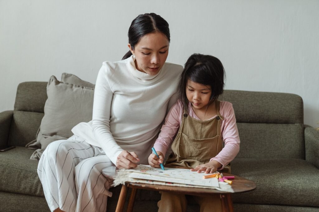 Woman sitting beside a young girl coloring in a book at a low table in front of a sofa.