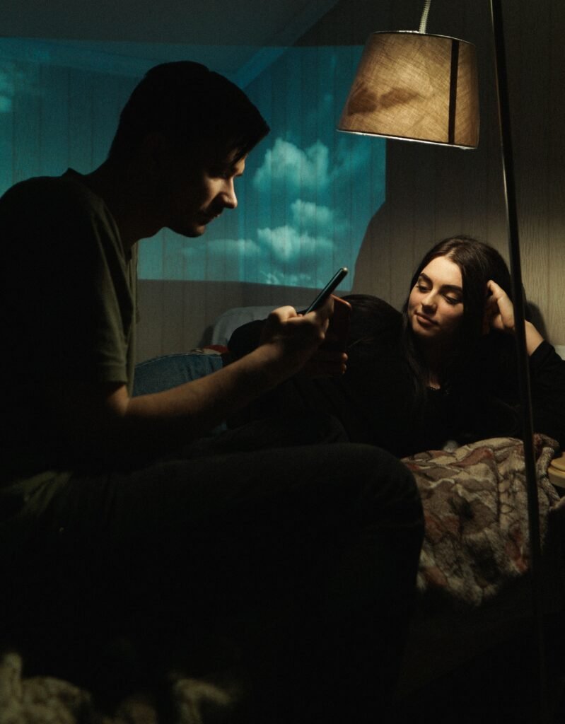 “A man holding a phone beside a woman seated on a sofa under a lamp.”