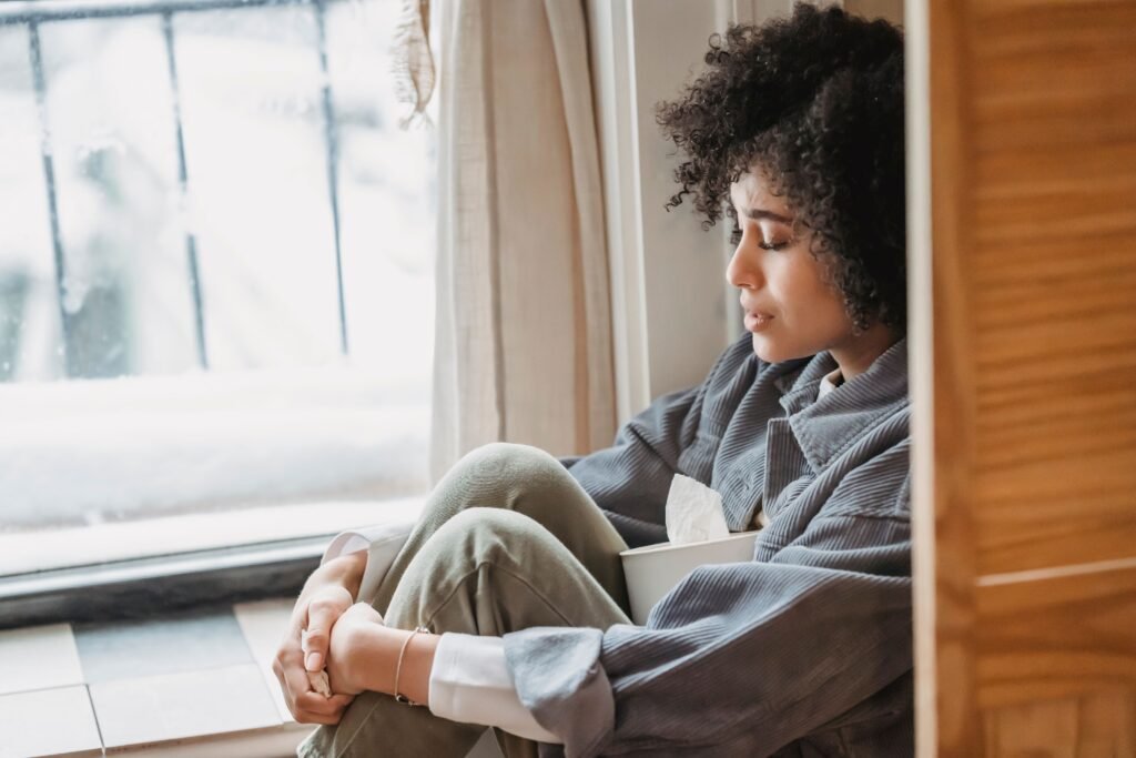 A woman sits by a window with a box of tissues, looking down.