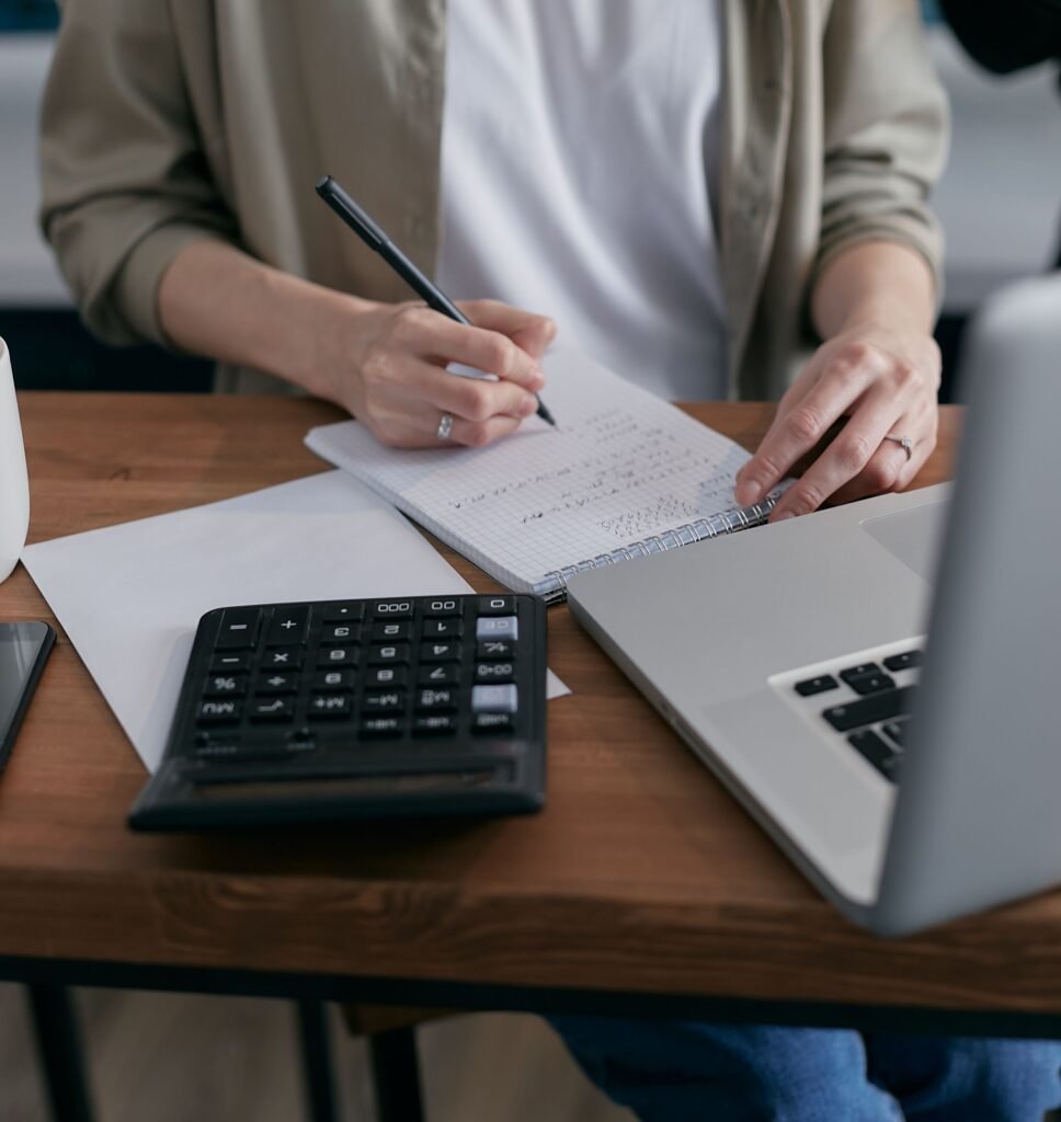 Person writing financial notes in a notebook at a wooden desk with a calculator and laptop, organizing household expenses.