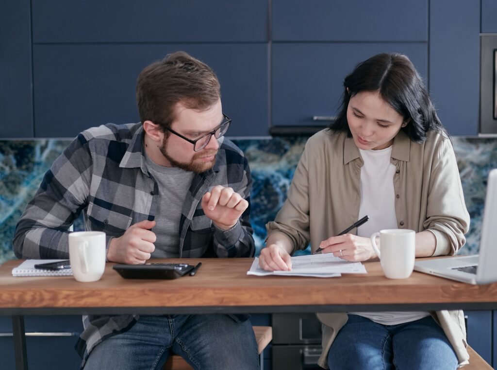Couple sitting together at a kitchen table, reviewing paperwork and discussing household finances with a calculator and laptop.